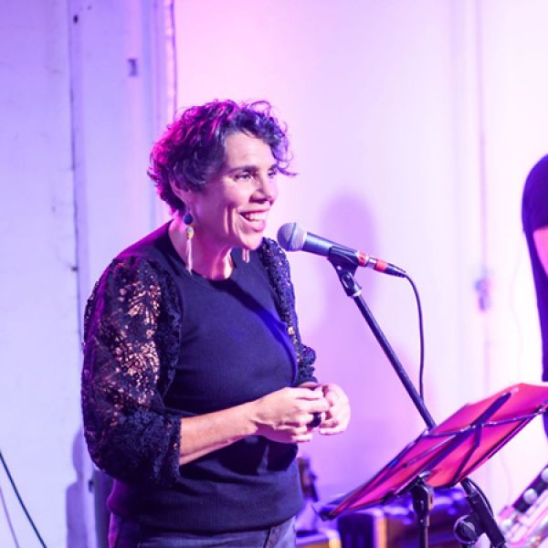 A person stands at a microphone beside a music stand, speaking or performing on a softly lit stage with purple and pink lighting.