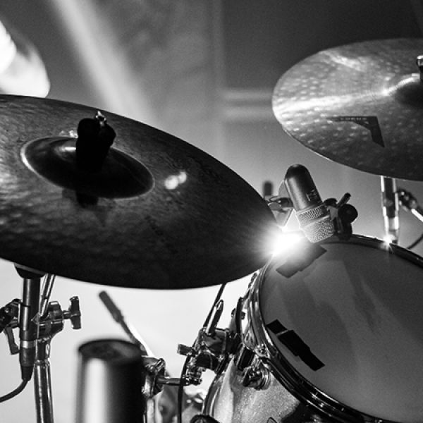 Black-and-white close-up of a drum kit with cymbals and a microphone, illuminated by dramatic stage lighting during a live percussion performance.