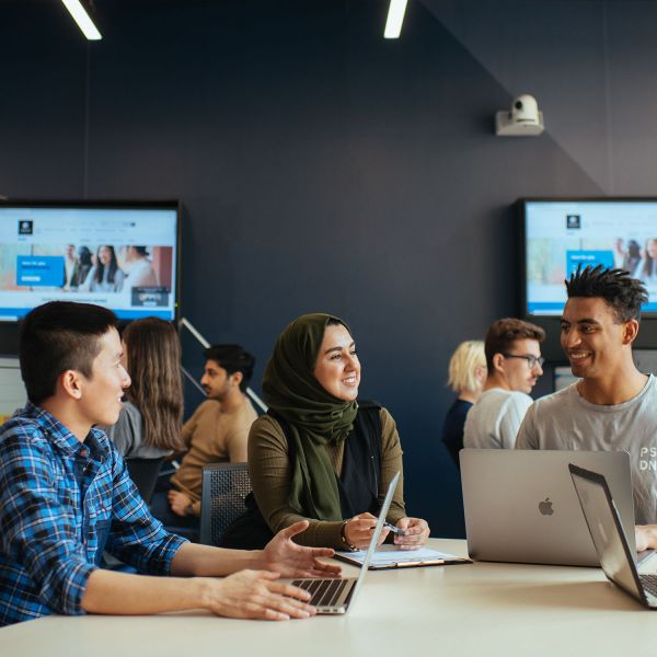 A diverse group of students sit at a table, with laptops in front of them, chatting and laughing.. 2022 QS World Rankings