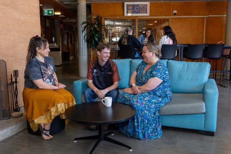 Three people sit on lounges around a small table, talking in an indoor university space.
