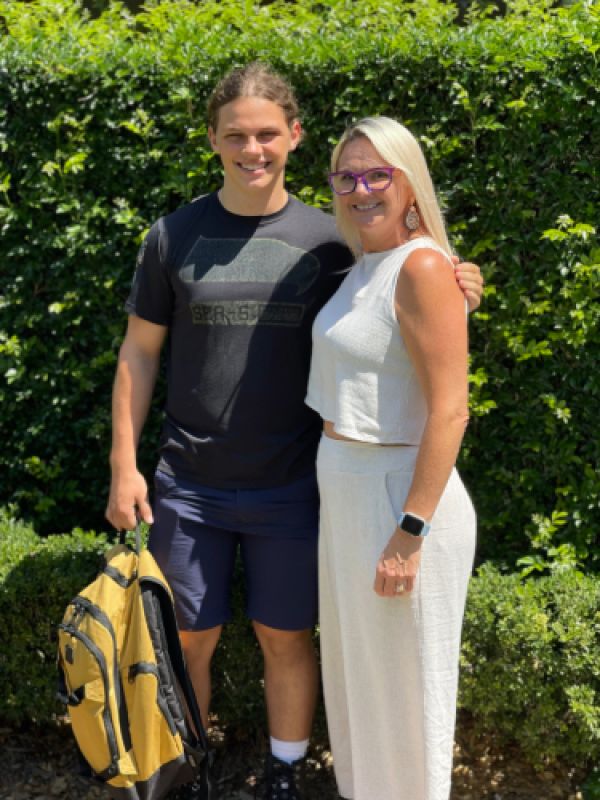 Letitia stands outdoors with her son Joe on a sunny day, smiling as they pose together in front of greenery, with Joe holding a backpack