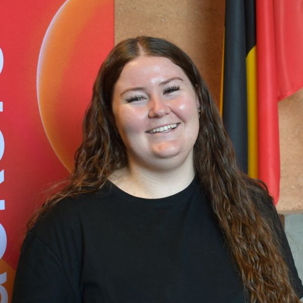 Hannah Cooper standing in a black shirt at the University of Newcastle MOU event, with CareerTrackers branding and an Aboriginal flag visible in the background.. New partnership to bolster Indigenous Careers