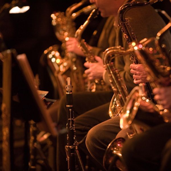 Close-up of several seated musicians in warm stage lighting playing saxophones and a clarinet, with shiny brass instruments and a music stand visible in the foreground.