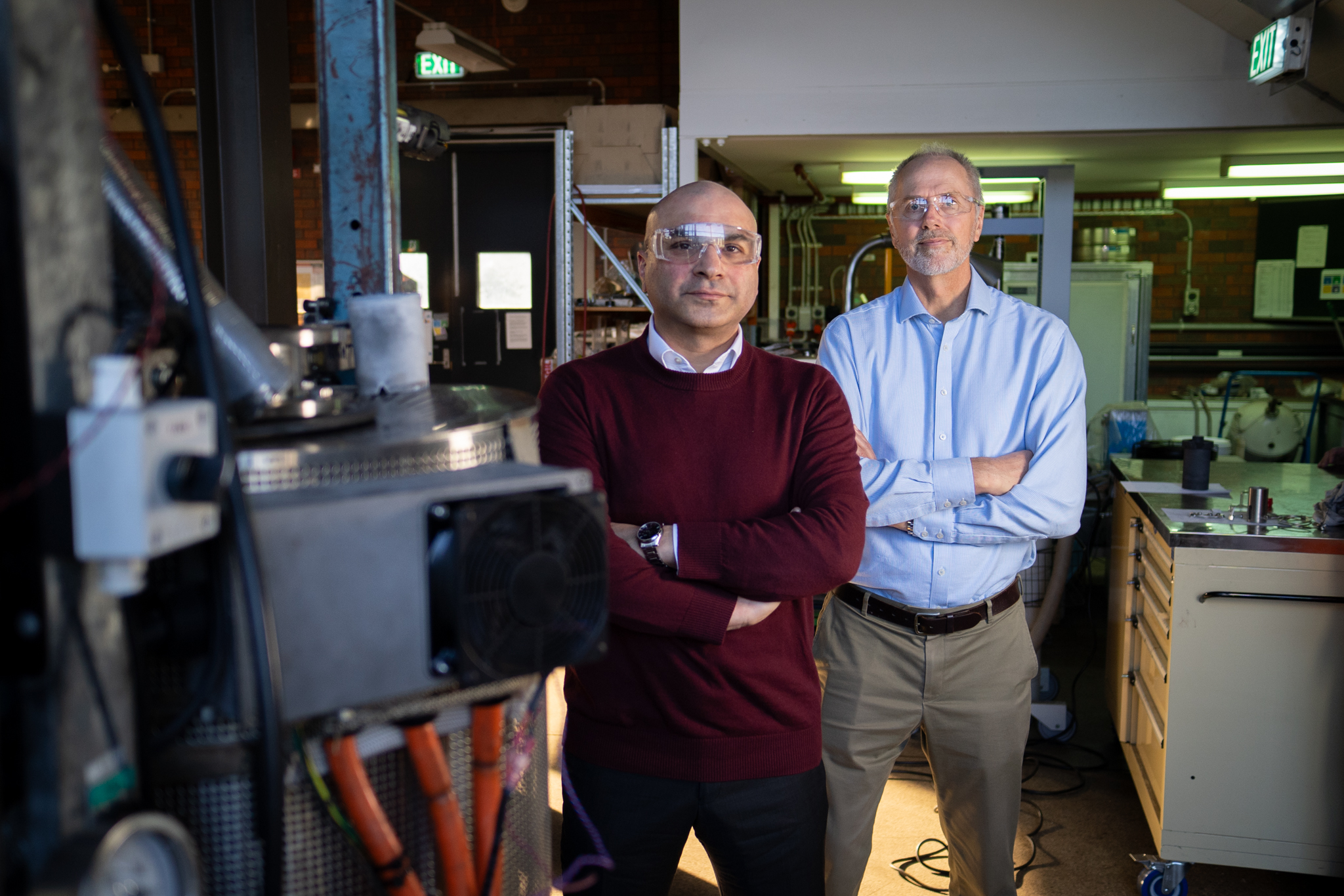 Two men stand in a lab looking at the camera. They both have their arms crossed and are wearing safety glasses. They are standing behind some machinery