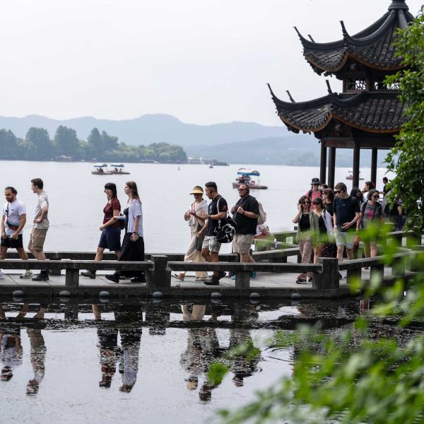Participants in the Ma & Morley China Immersion Tour walk along a lakeside boardwalk beside a traditional Chinese pavilion, with boats and distant hills visible across the lake. Approaching a decade of difference.