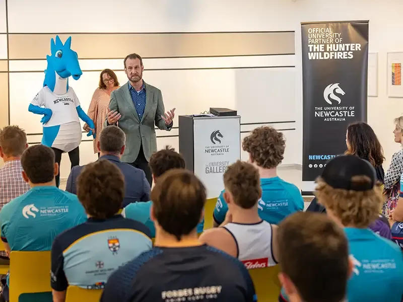 A speaker addresses an audience seated in an auditorium, with the blue University of Newcastle seahorse mascot standing in the background.