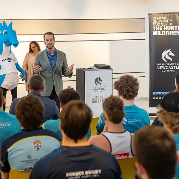 A speaker addresses an audience seated in an auditorium, with the blue University of Newcastle seahorse mascot standing in the background.
