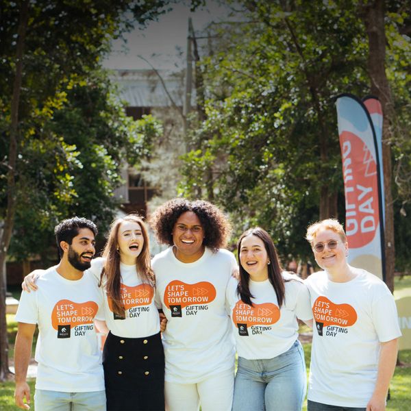 3 male and 2 female students all in white Gifting Day T-Shirts looking to camera smiling standing side by side with their arms around each other's shoulders.