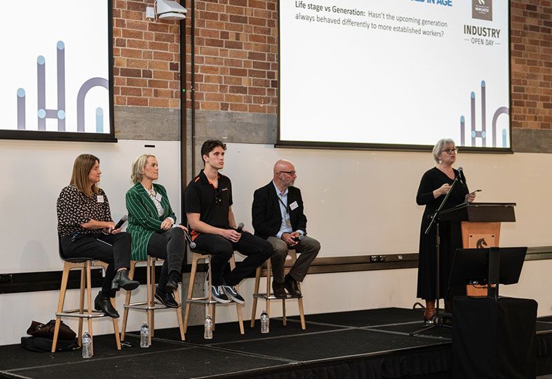 Four panellists sitting on stools on a stage, and one woman standing at a podium