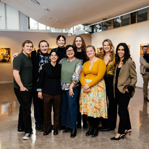 Posed group picture of students, staff and community members positioned in front of the 50 Years, Fifty Stories Exhibition at the University Gallery. . Celebrating 50 years of Open Foundation