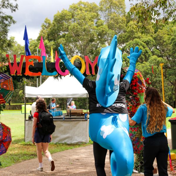 The University of Newcastle mascot, Hunter the Hippocampus - a big blue mascot - raises their hands as they walk through a big Welcome archway