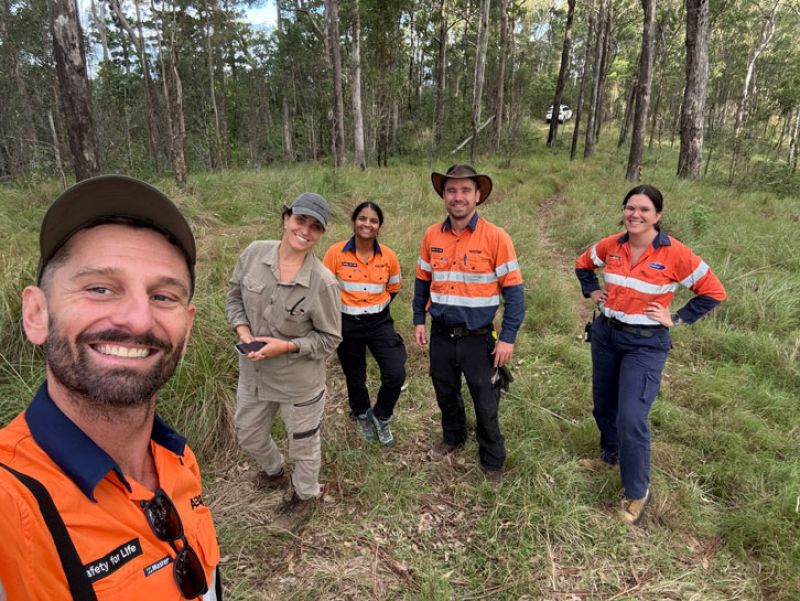 Five Ecologists, including Sonny, in uniforms and hats, posing in a wooded area, smiling at the camera.