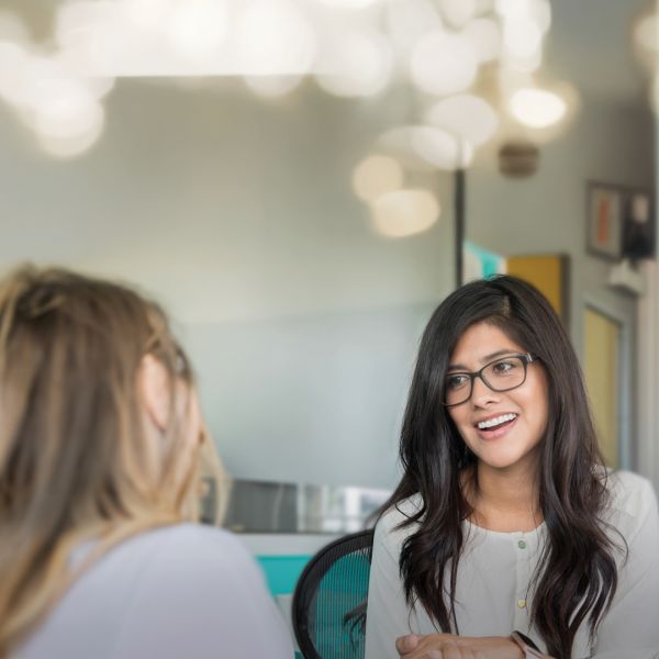 Woman facing camera, smiling to woman facing away from camera. In an indoor office setting. - Master of Clinical Psychology and Master of Professional Psychology Webinar