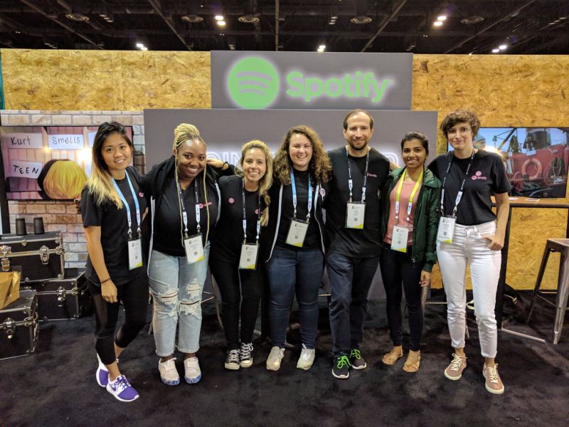 Chris stands in a row alongside 6 female colleagues at the Grace Hopper conference. They all wear black t-shirts with Spotify logos standing together, smiling in front of a Spotify exhibition booth with a wood-paneled background.