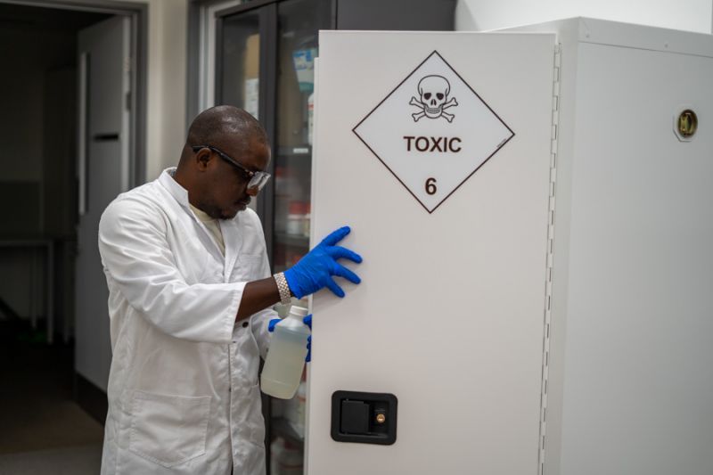 A scientist wearing gloves and protective glasses, puts a sample bottle into a cupboard with a toxic warning sign