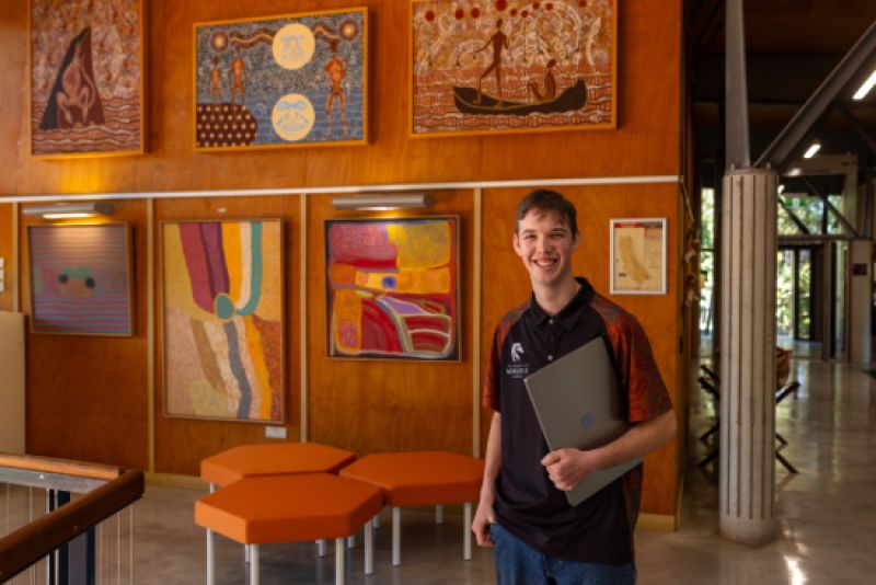 A young man stands inside a university building, with colourful, Aboriginal artwork displayed on the wall behind him.