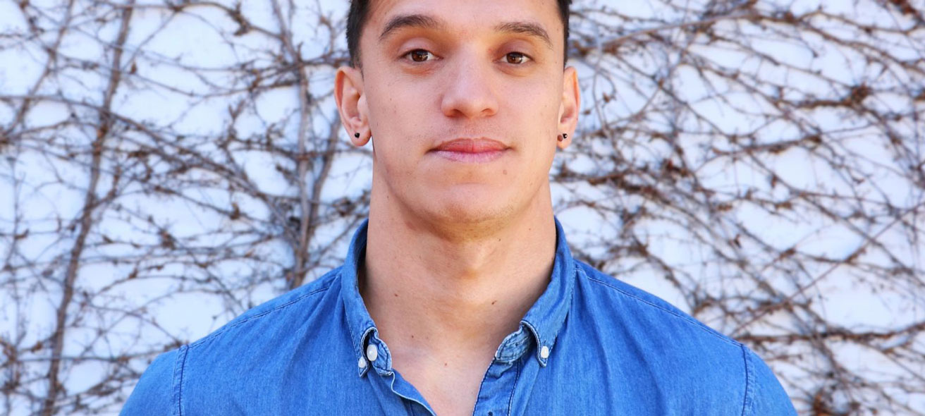 Tualagi is wearing a blue shirt and looking at the camera. Standing in front of a generic / unknown background.