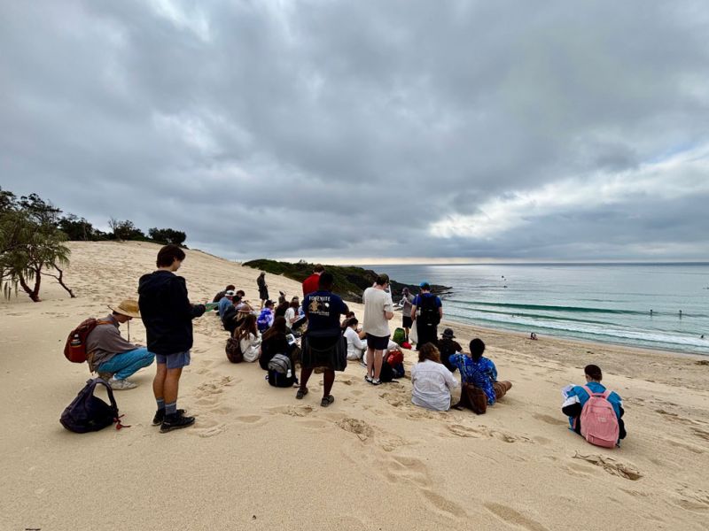 Students and staff sit and stand on sand near the ocean.