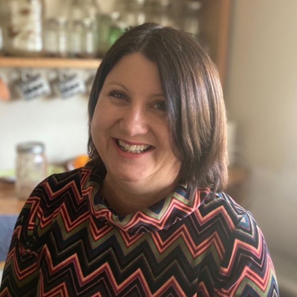 A close-up portrait shows a person smiling at the camera, with short dark hair and wearing a colorful zigzag-patterned top, standing in a softly lit kitchen with shelves and jars in the background. Provide your feedback on BizChat