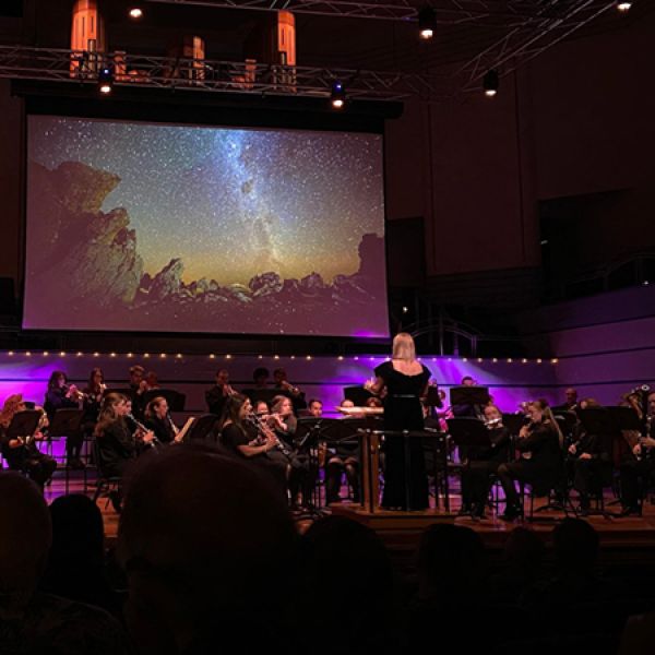 A conductor leads a seated orchestra on a lit stage while a large screen above displays a starry night sky over rocky terrain.