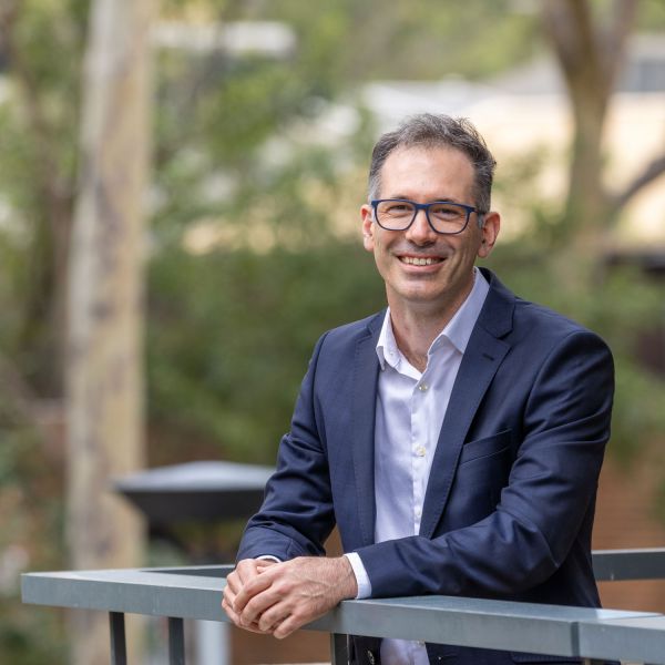 Lachlan wearing a navy suit jacket and glasses, leaning on a railing outdoors, with trees and greenery softly blurred in the background. Why I donate – Lachlan King