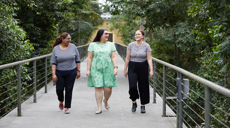 Three people smiling and talking while walking over a bridge surrounded by foliage.