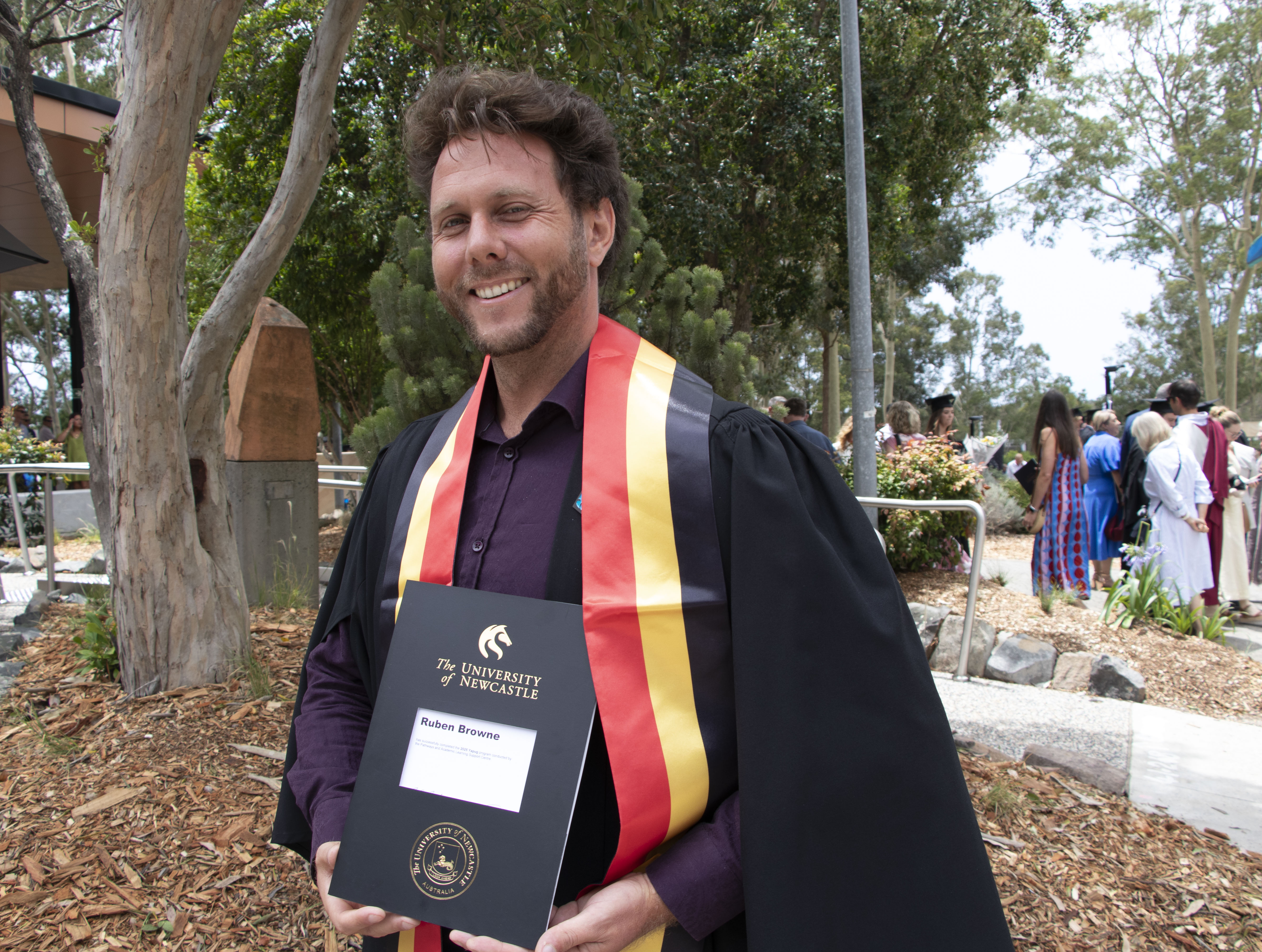 A man stands in his graduation garb proudly holding his degree. He is smiling and has a black, red and yellow sash on. 