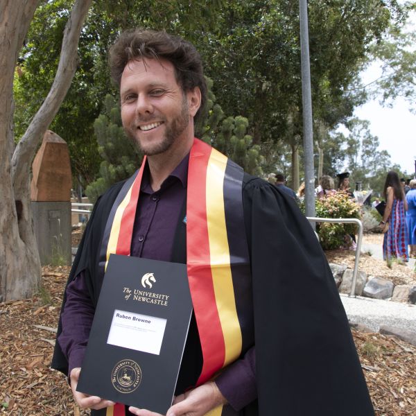 A man stands in his graduation garb proudly holding his degree. He is smiling and has a black, red and yellow sash on. 