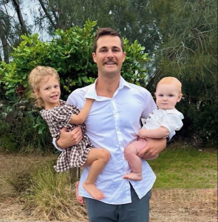 A man stands proudly smiling holding a little girl on his left hip and small baby boy on his right hip. All are very happy. 