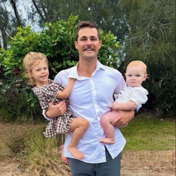 A man stands proudly smiling holding a little girl on his left hip and small baby boy on his right hip. All are very happy. 