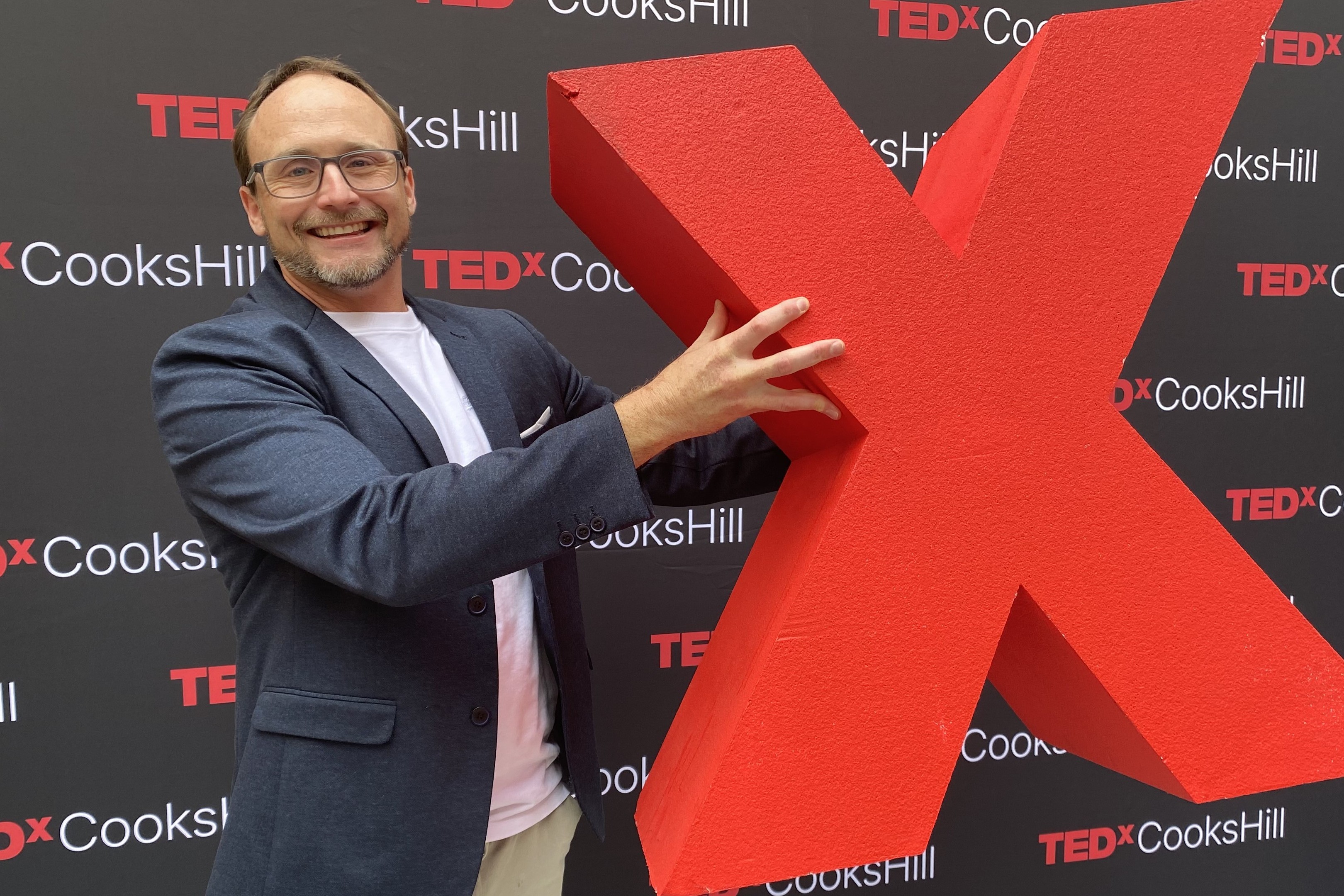 Dr Daniel Beard holding an oversized foam letter X in front of the TEDx Cooks Hill banner