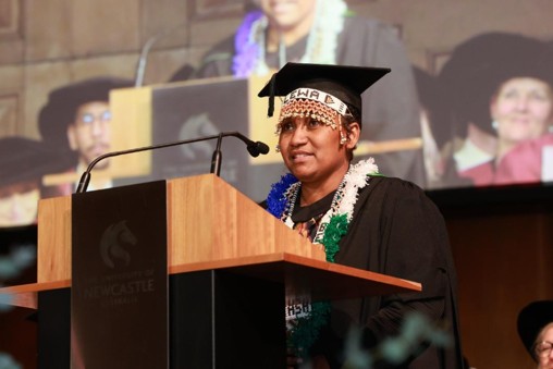 Women in graduation cap and gown and traditional Pacific attire stands behind lectern to deliver speech