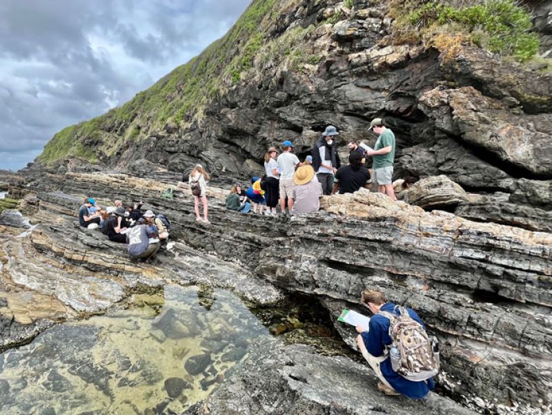 Students stand on rock, observing the environment and noting down items on clipboards.