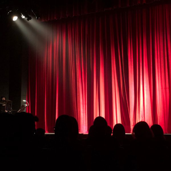 Red stage curtains glow under a spotlight while the silhouettes of audience members sit in the foreground.