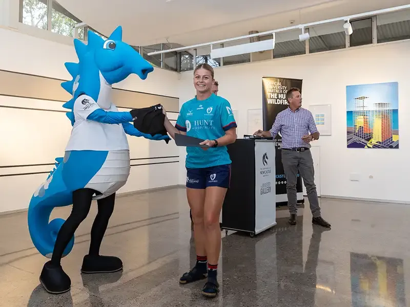 The blue University of Newcastle seahorse mascot hands an item to an athlete at a presentation.