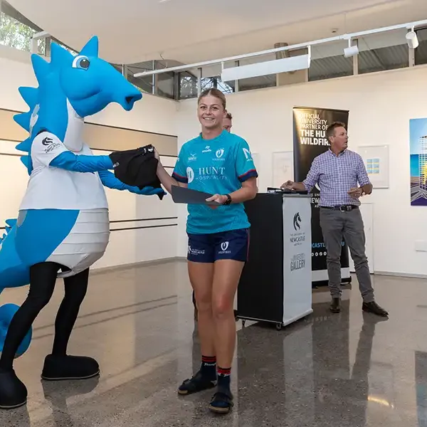 The blue University of Newcastle seahorse mascot hands an item to an athlete at a presentation.