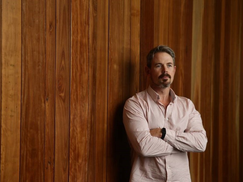 Nathan Barlett standing in front of a brown wooden wall with his arms folded