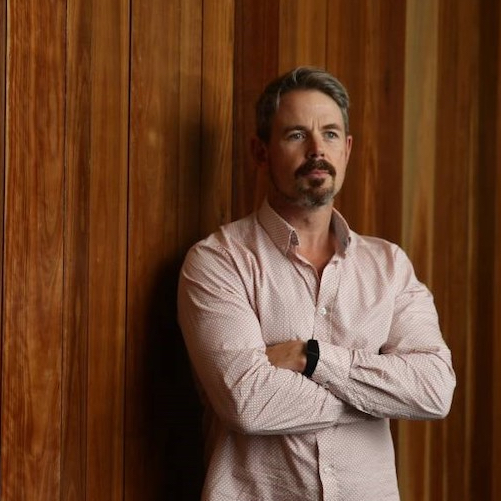 Nathan Barlett standing with his arms folded in front of a wooden wall