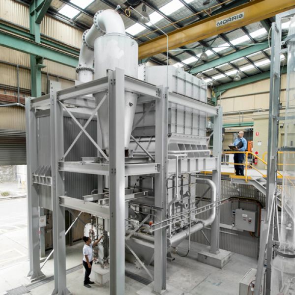 Image shows huge grey machinery, a person in a white shirt stands at the bottom and a person in a blue shirt with a clipboard is off the the right at the top of a staircase